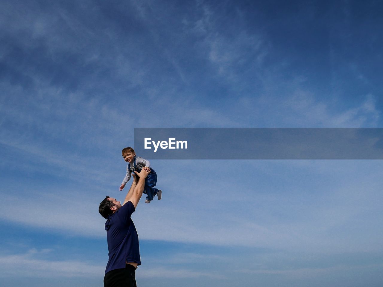 Low angle view of father holding cute baby son while standing against blue sky
