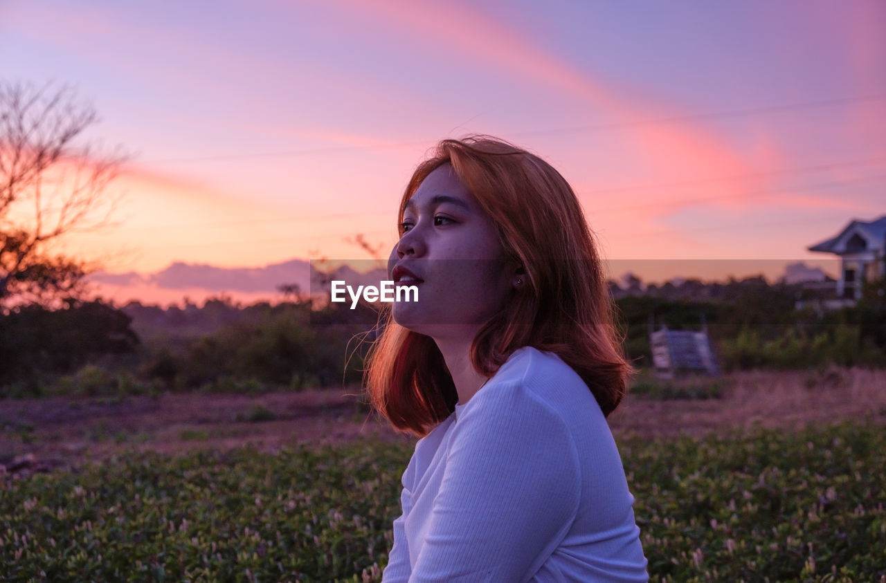 Portrait of young woman sitting on field against sky during sunset