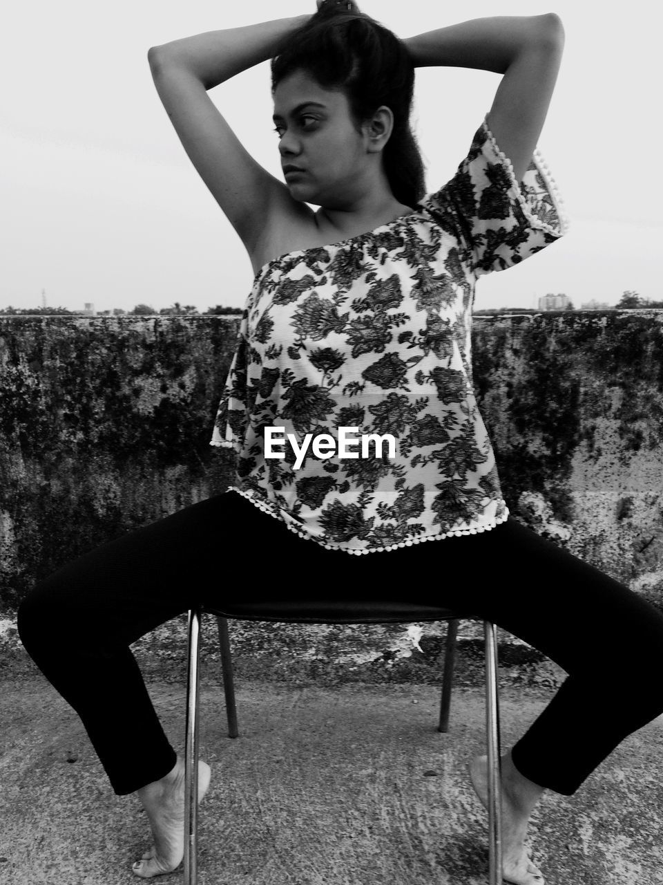 Young woman with hands in hair sitting on chair against sky