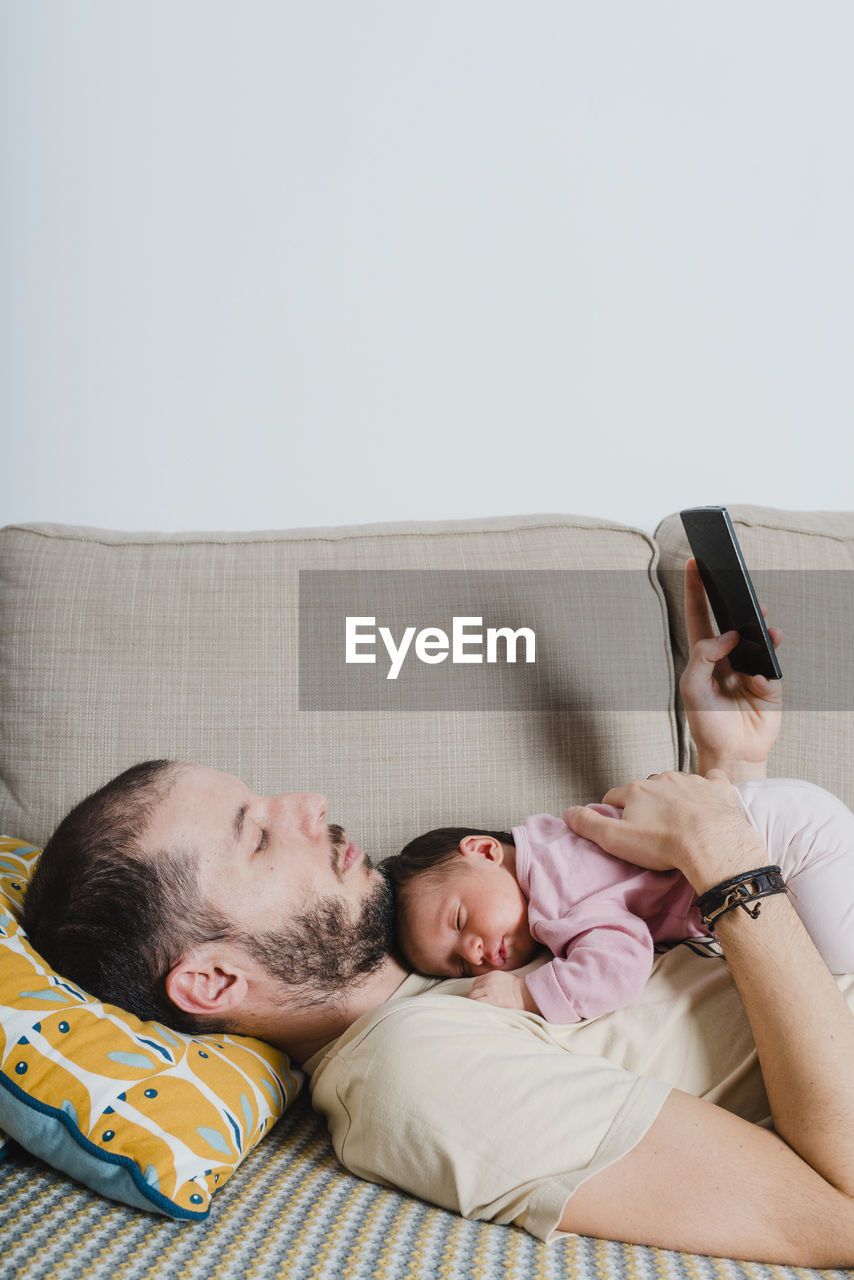 Father lying on the couch with newborn baby girl using smartphone