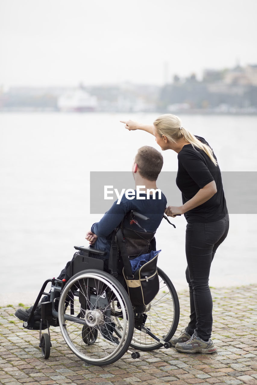 Caretaker showing something to disabled man on wheelchair by lake