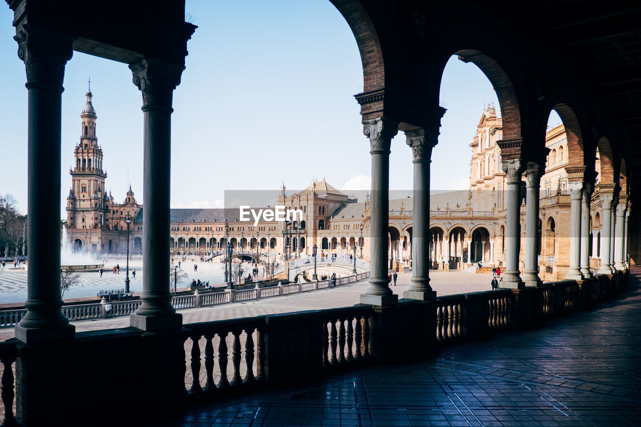 Historic buildings at plaza de espana seen through colonnade against blue sky