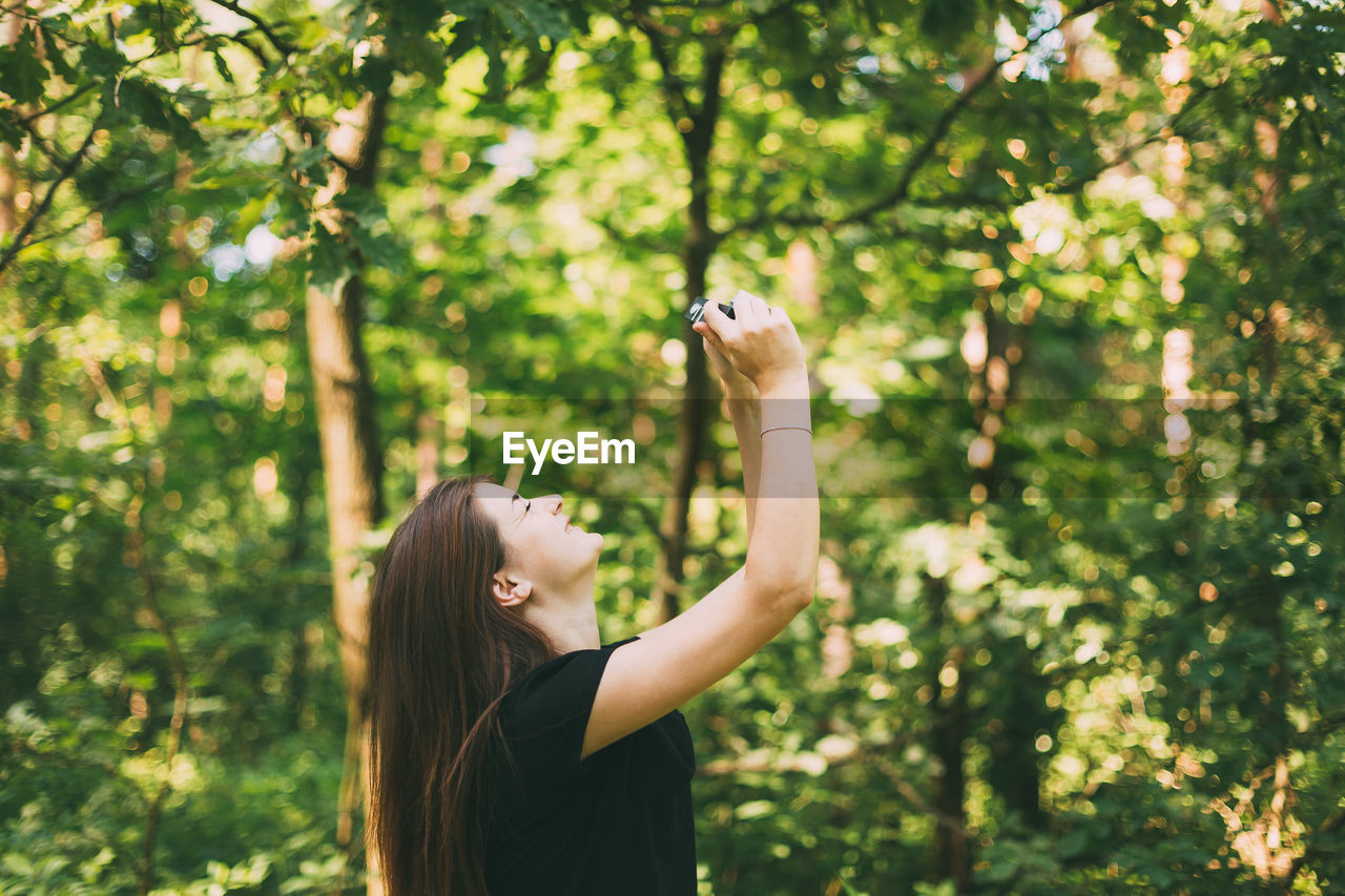 Side view of young woman photographing through camera in forest