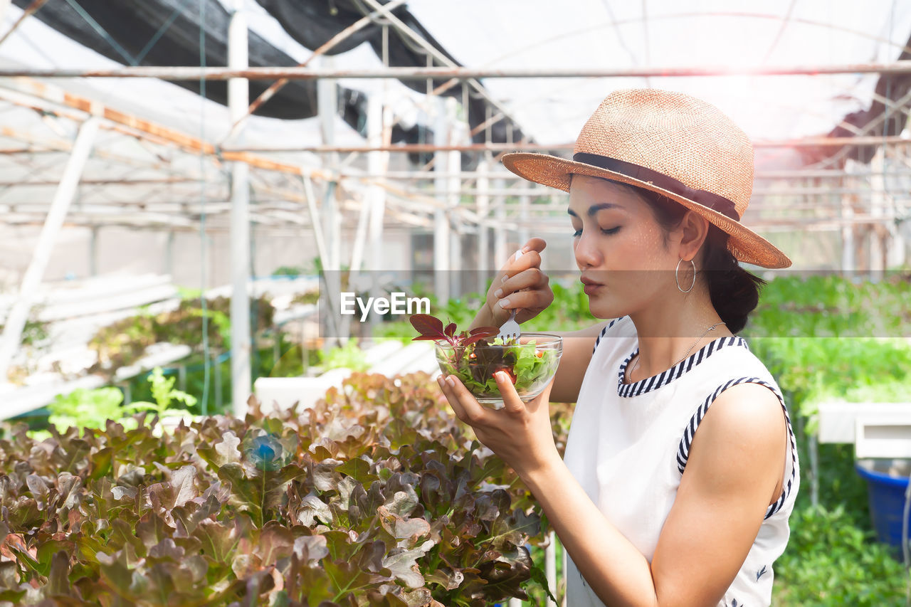 Woman wearing hat while holding salad bowl at greenhouse