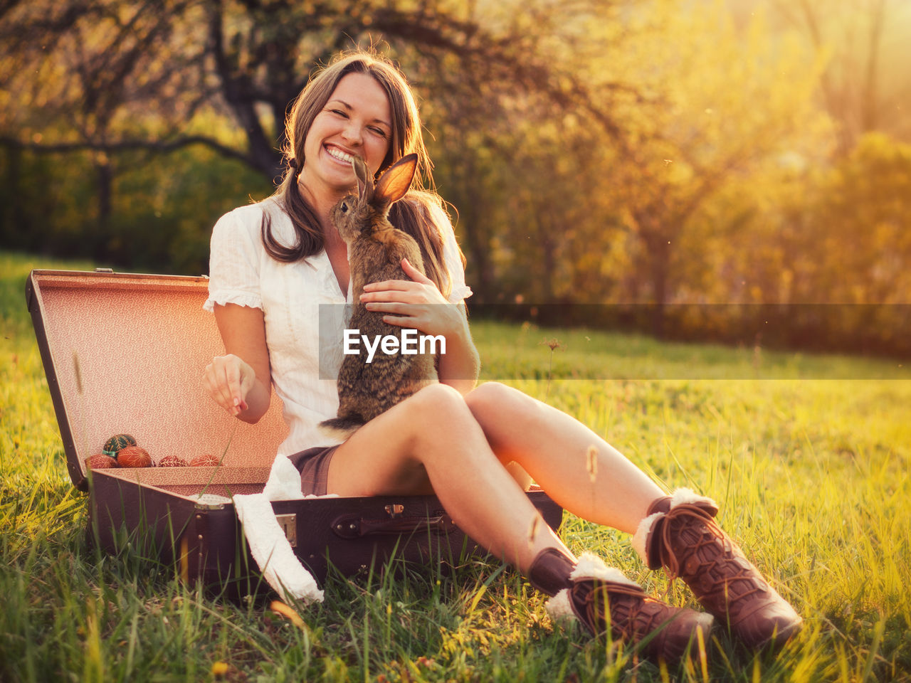 Portrait of smiling woman with rabbit sitting in box against trees at park