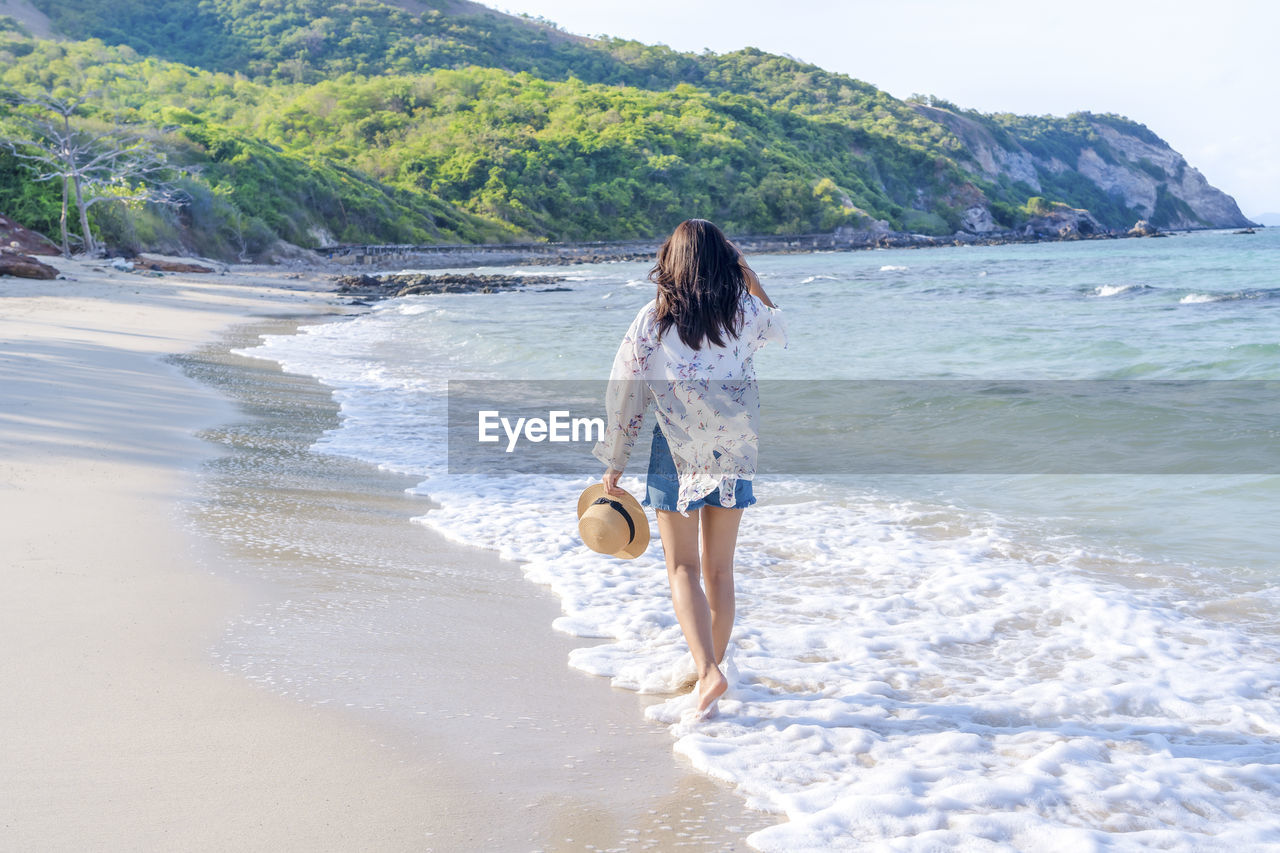 Asian woman holding back straw hat walking on the beach during weekend summer vacation 