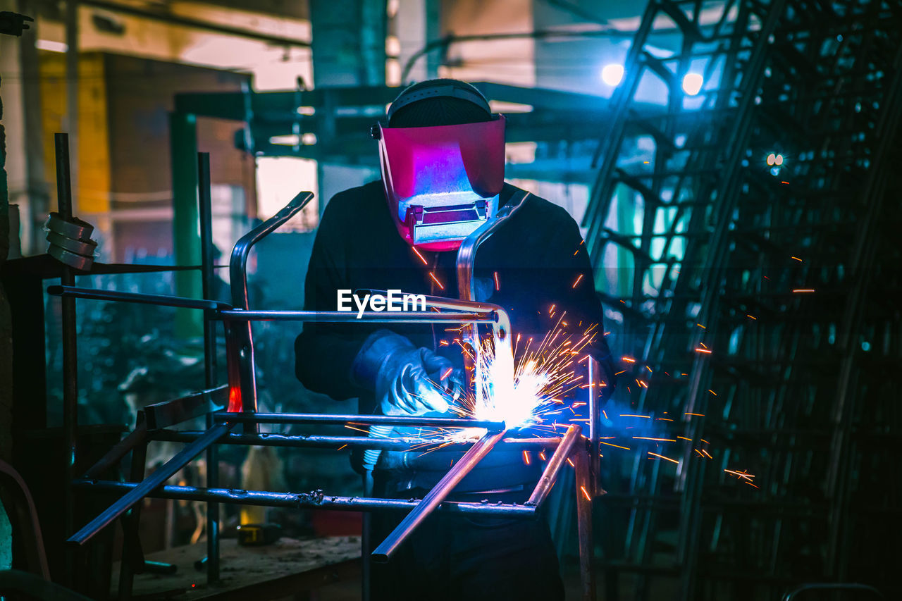 Welder in production is engaged in the manufacture of products. in a mask, in gloves, sparks fly
