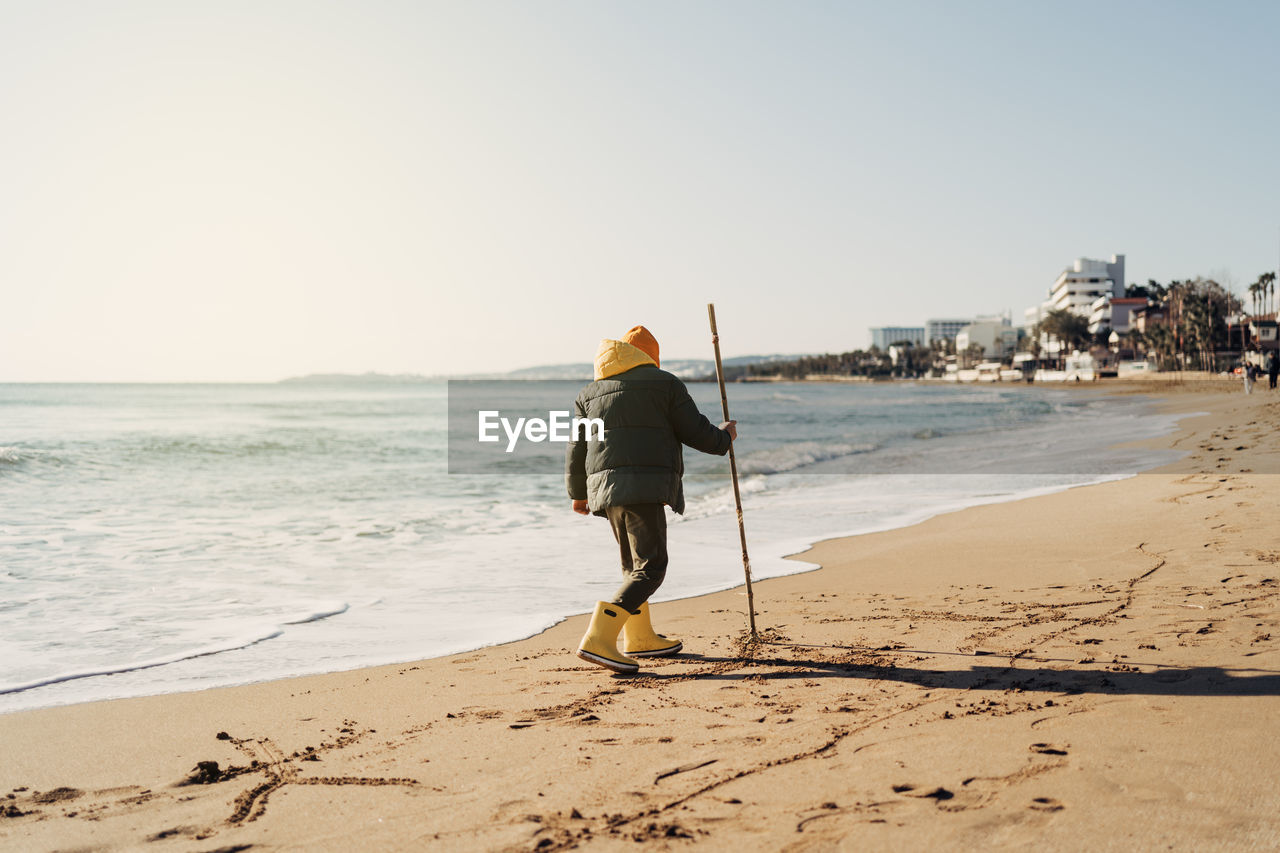 Boy in yellow rubber boots playing with water and sand at the beach. child kid at autumn winter sea.