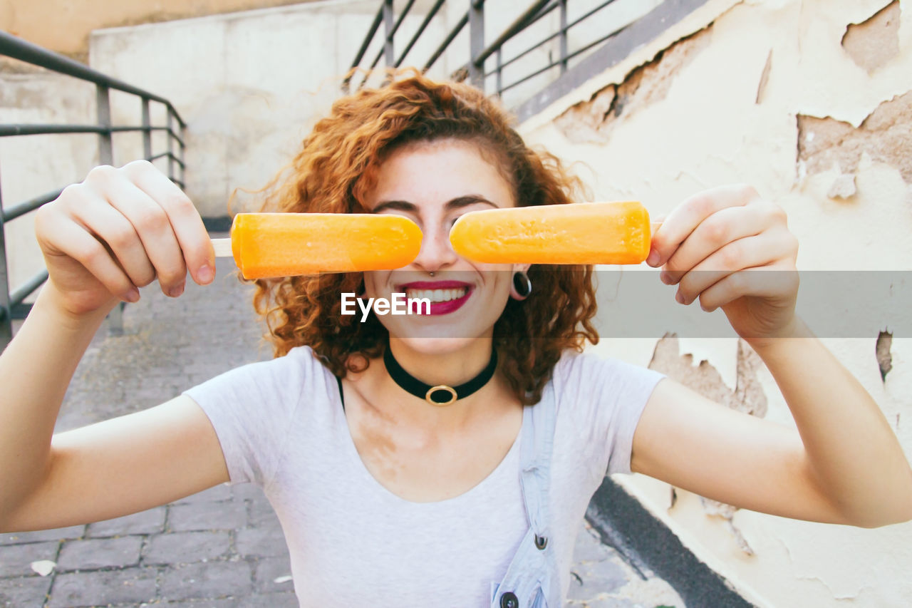 Close-up of woman smiling while holding orange popsicles on footpath