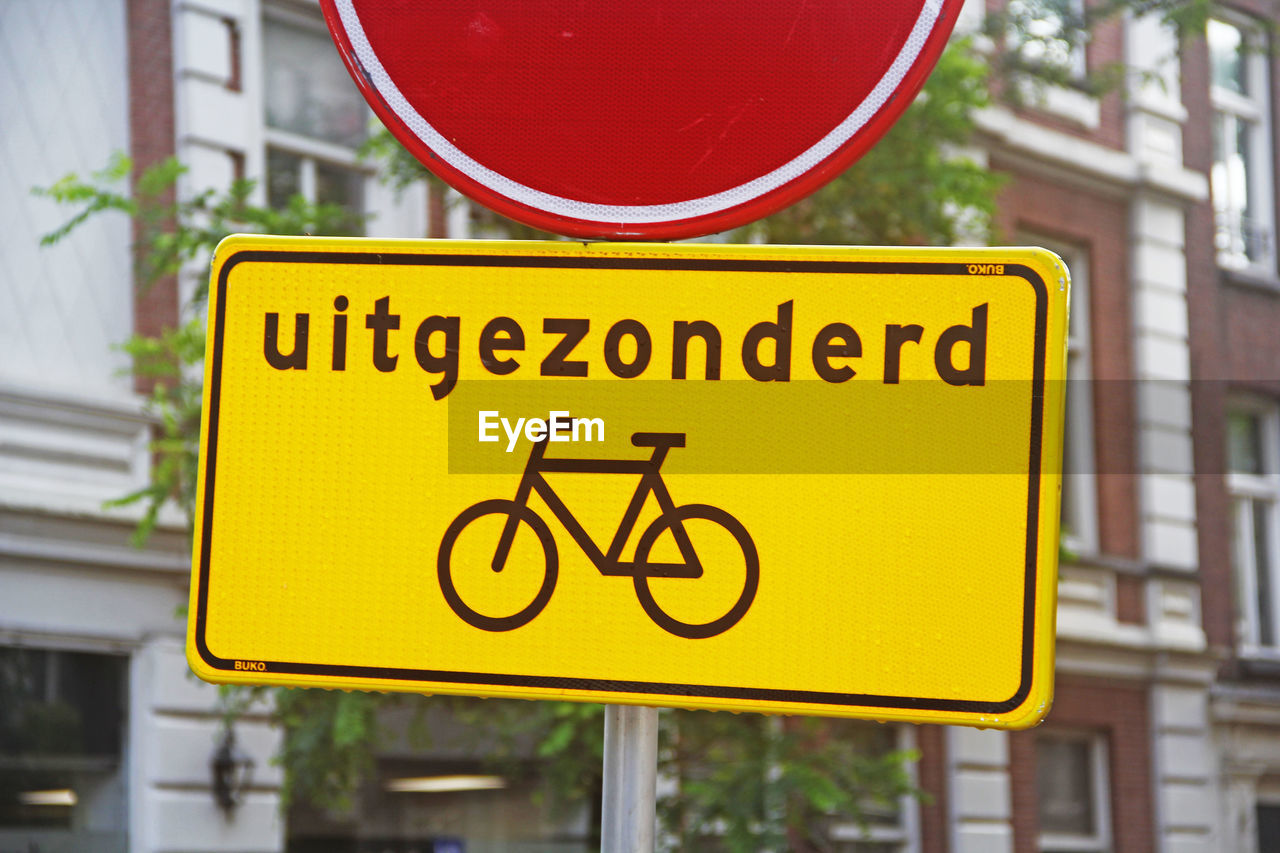 sign, communication, traffic sign, bicycle, road sign, city, text, yellow, signage, road, street sign, architecture, guidance, warning sign, no people, street, western script, transportation, day, lane, building exterior, red, outdoors, focus on foreground, symbol, information sign, city life, built structure, vehicle, city street