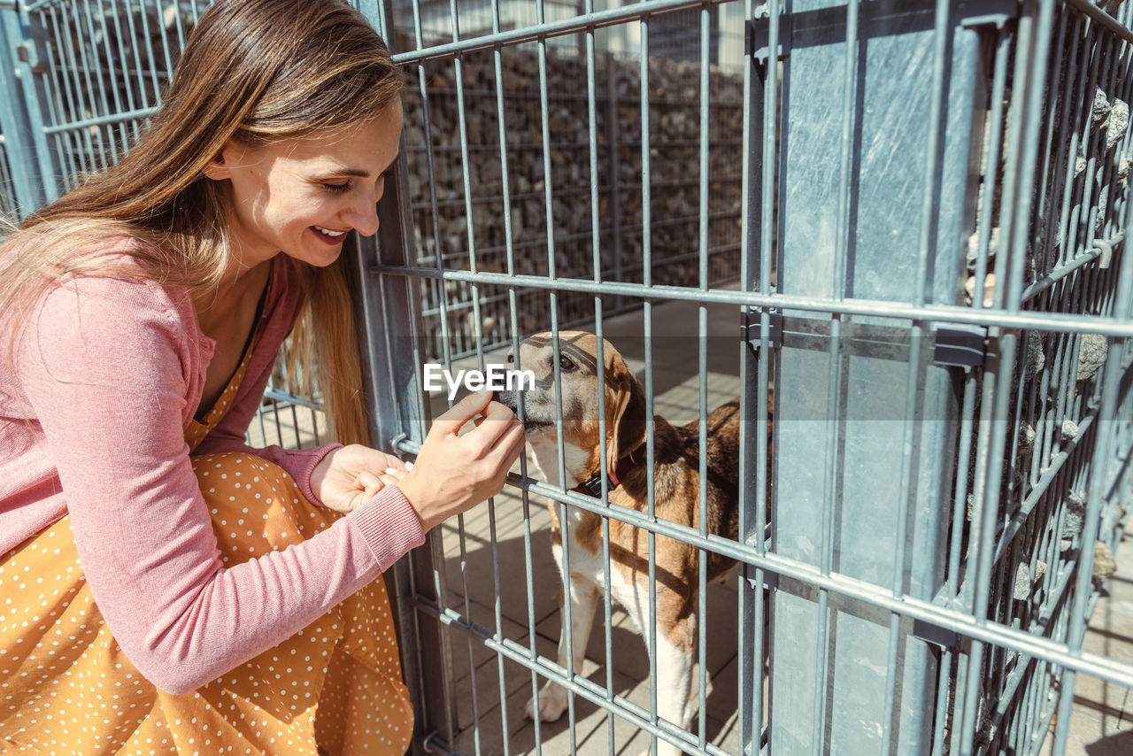 Smiling woman feeding food to dog in cage