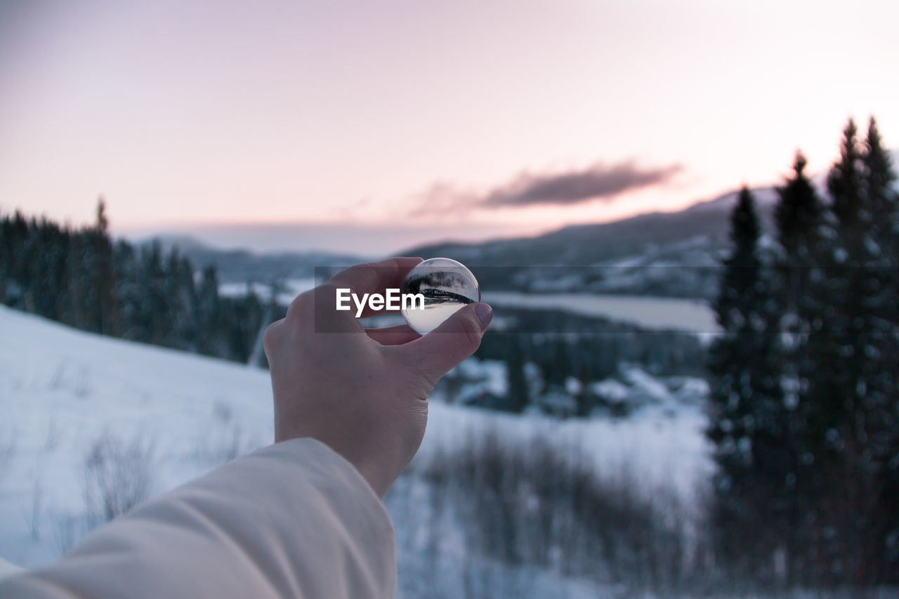 Cropped hand of person holding crystal ball against landscape during winter