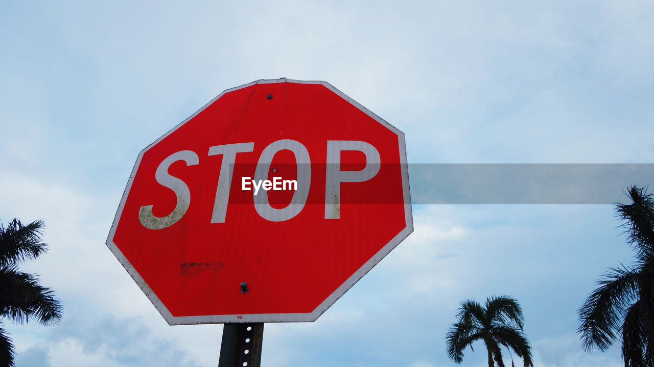 LOW ANGLE VIEW OF ROAD SIGN BY PALM TREES AGAINST SKY