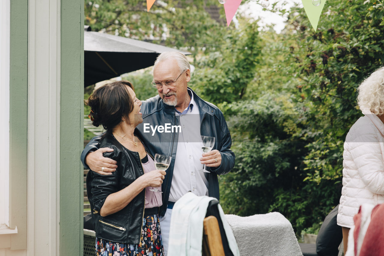 Smiling senior friends standing with arm around enjoying garden party at back yard