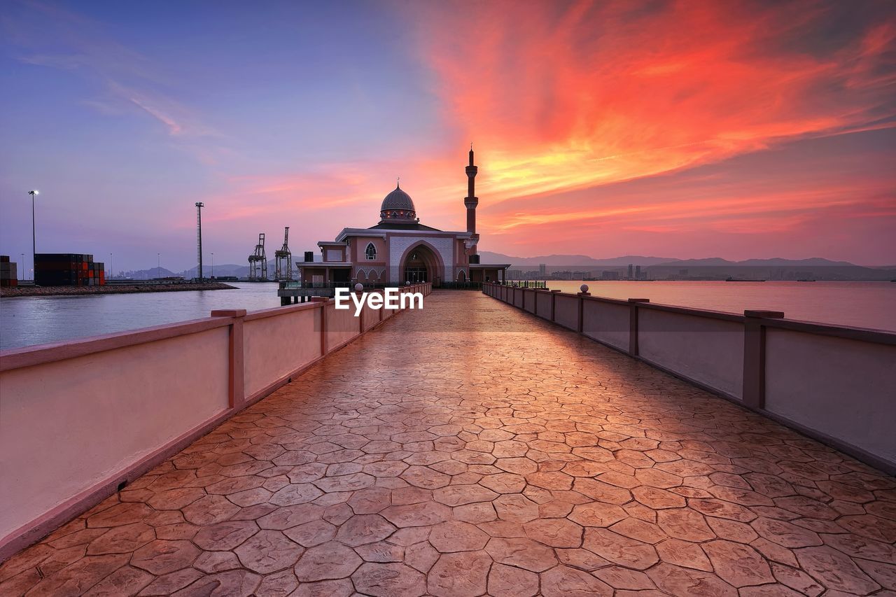 Walkway leading towards mosque by sea at sunset against sky