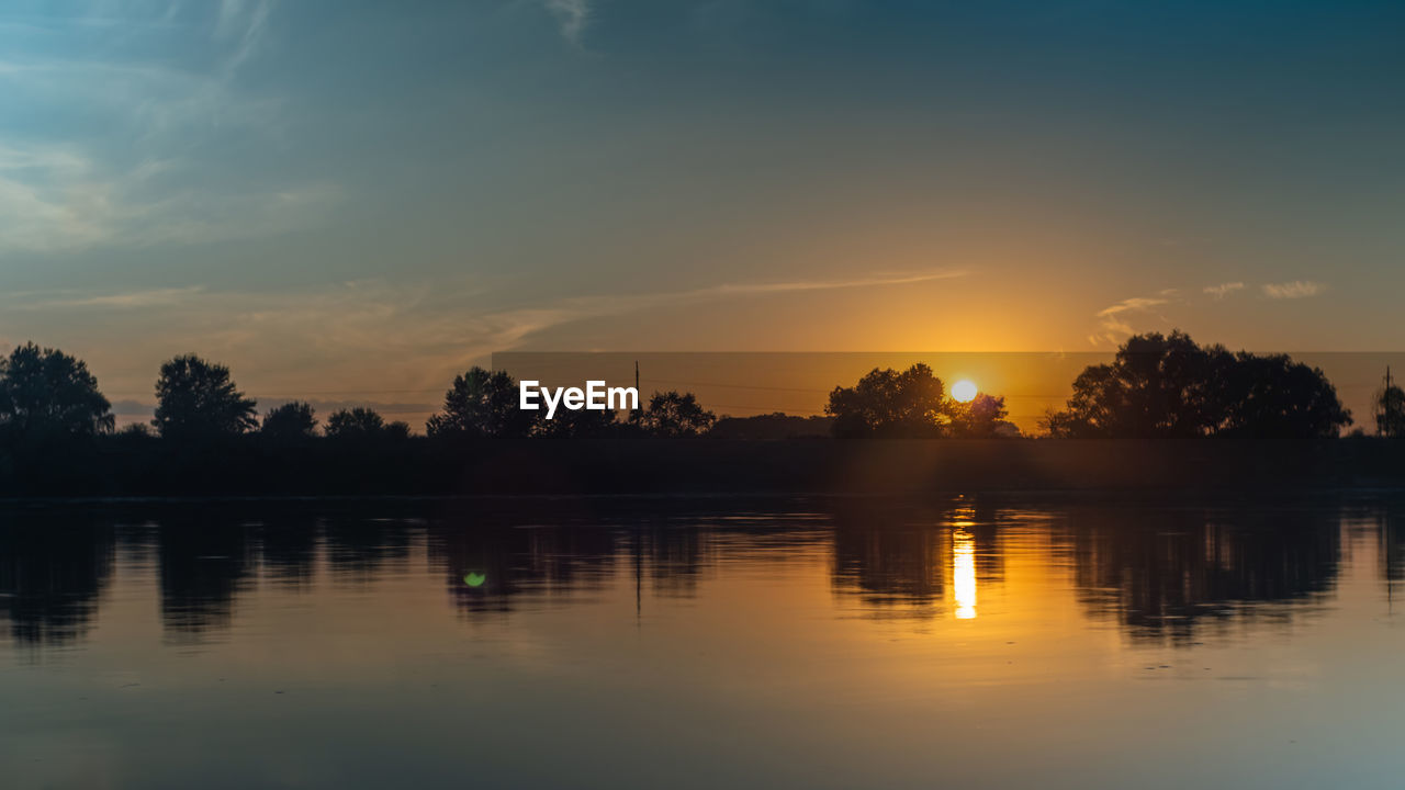 Summer sunset over the river. trees and clouds reflected in the water. orange ball of sun hiding