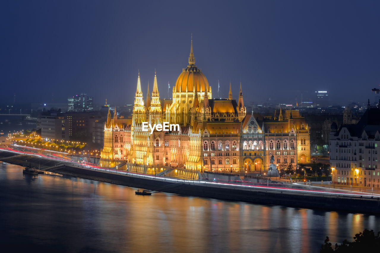 illuminated buildings by river against sky at night