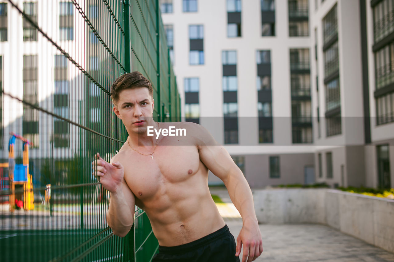 Portrait of muscular man standing by fence at park