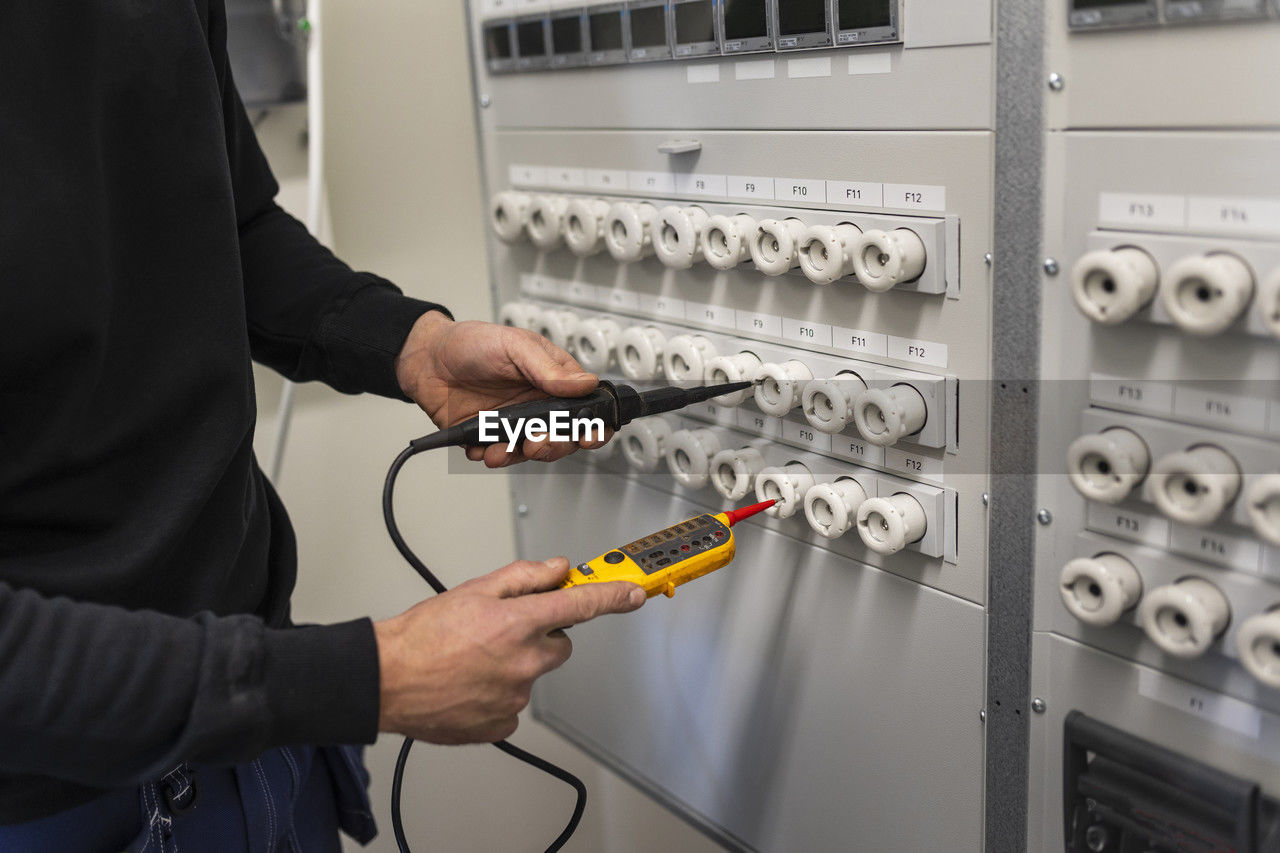 Midsection of male electrician with voltage tester repairing switchboard in meter room