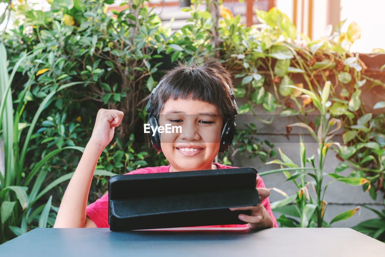 Asian cute boy wearing wireless headphones and smiling happily while playing games on the tablet.