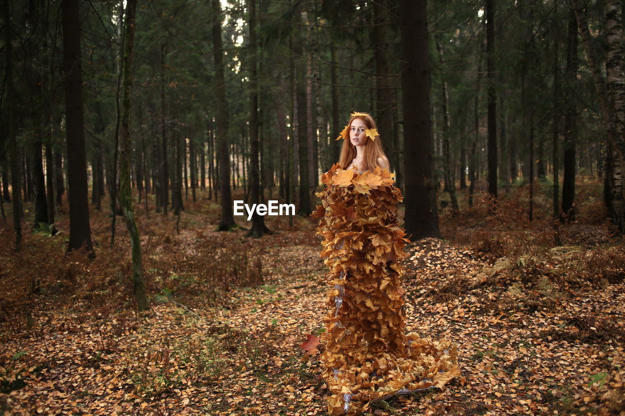 Portrait of young woman covered with leaves standing at forest during autumn