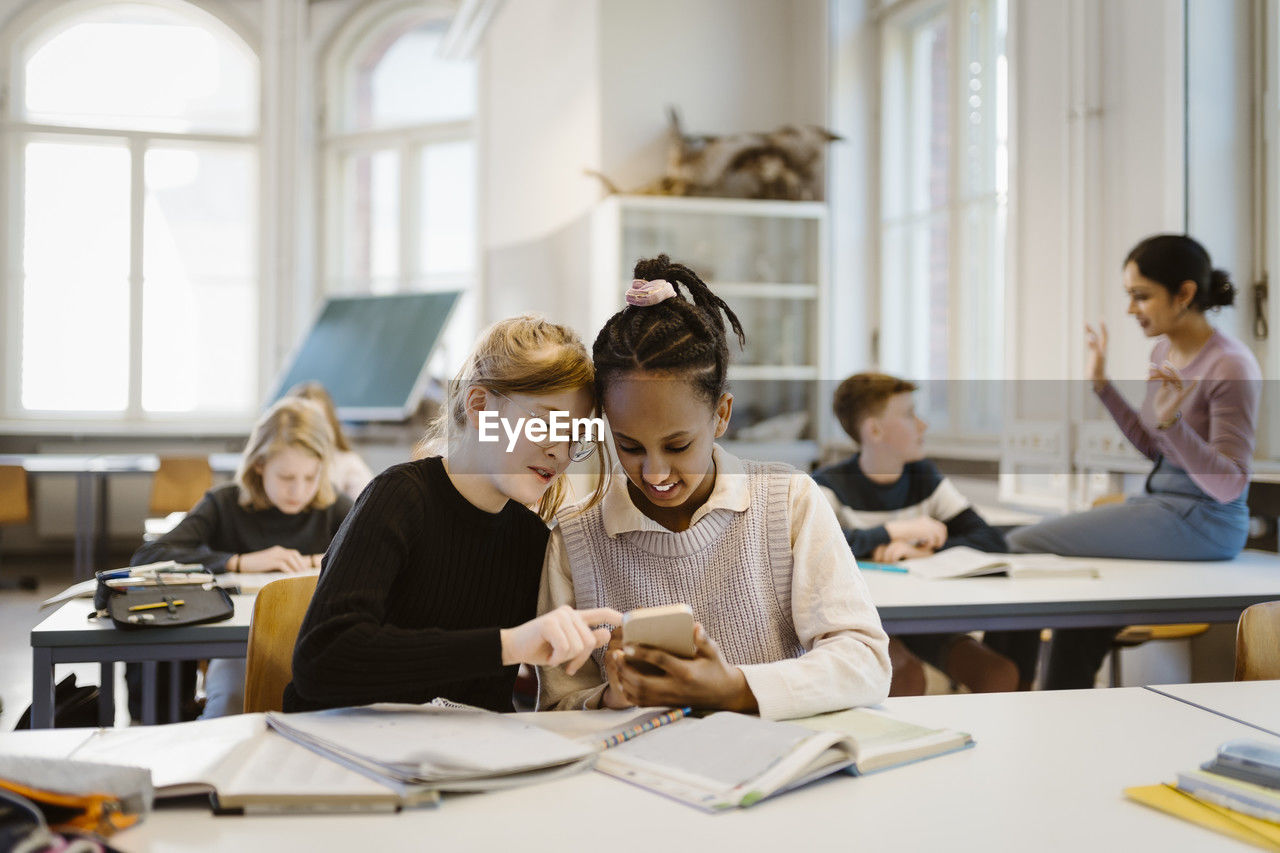 Curious schoolgirls sharing smart phone while sitting at desk in classroom