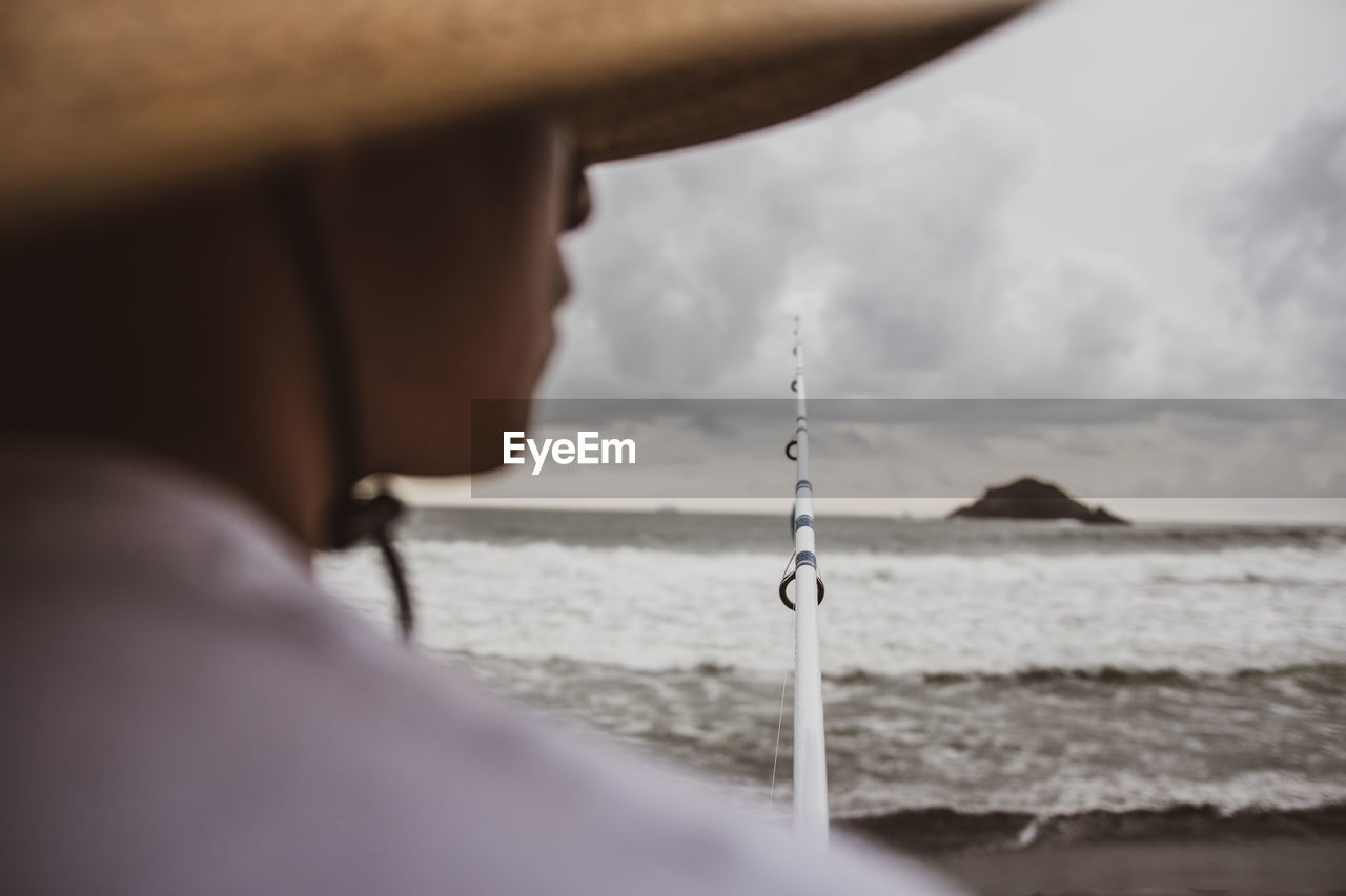 Side view of unrecognizable male in hat with fishing rod catching fish on sandy shore near waving sea