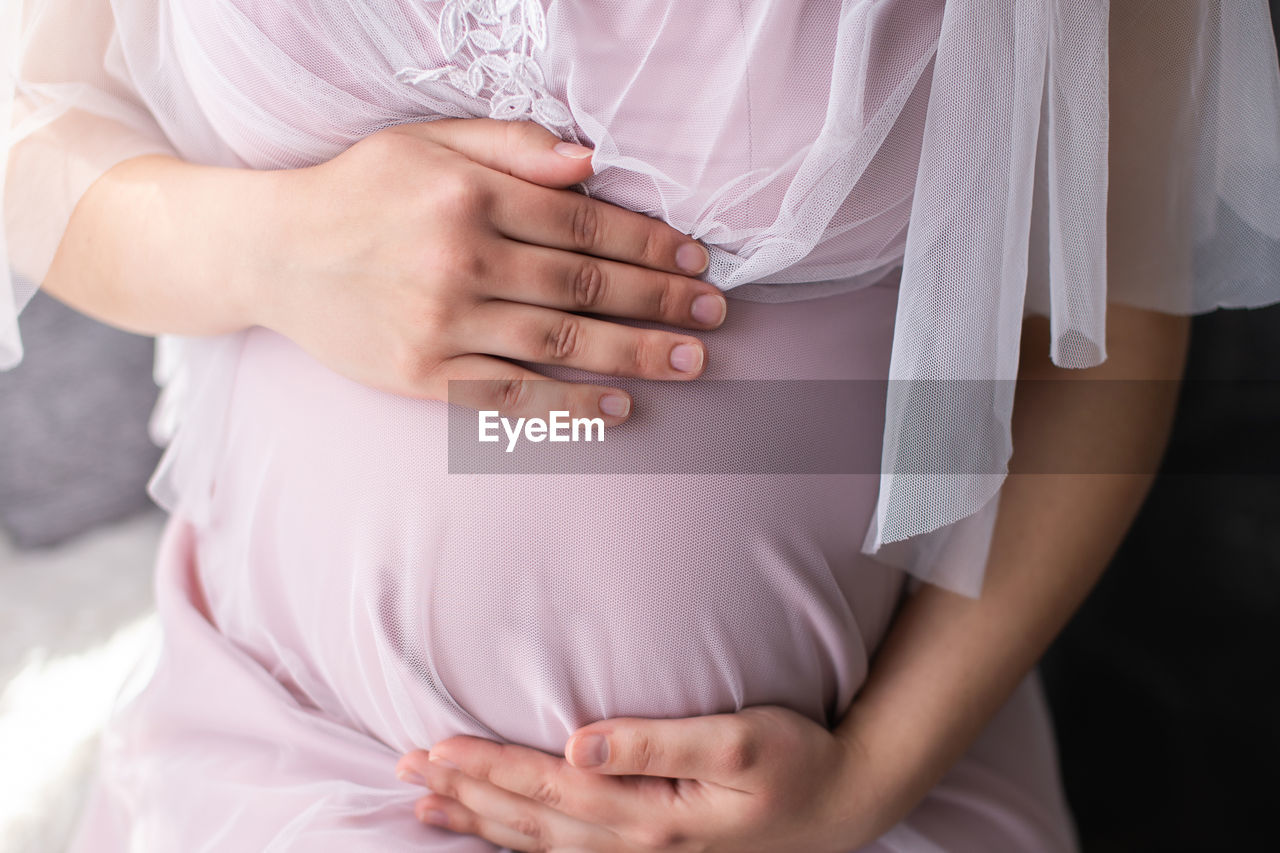 The belly of a pregnant woman in a pink dress holds her hands