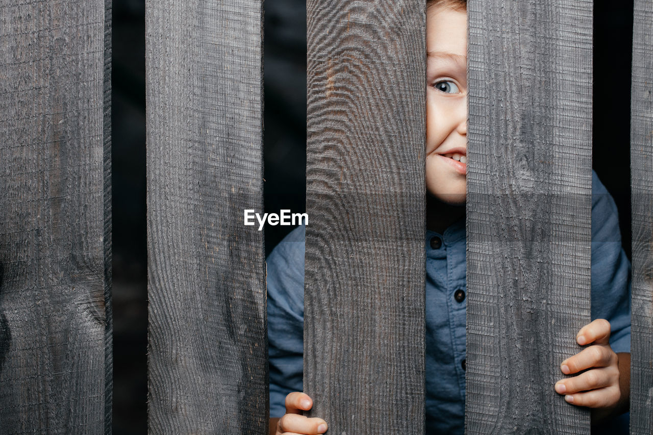 Happy smiling white boy looks out of the crack of a wooden fence. childish curiosity. espionage