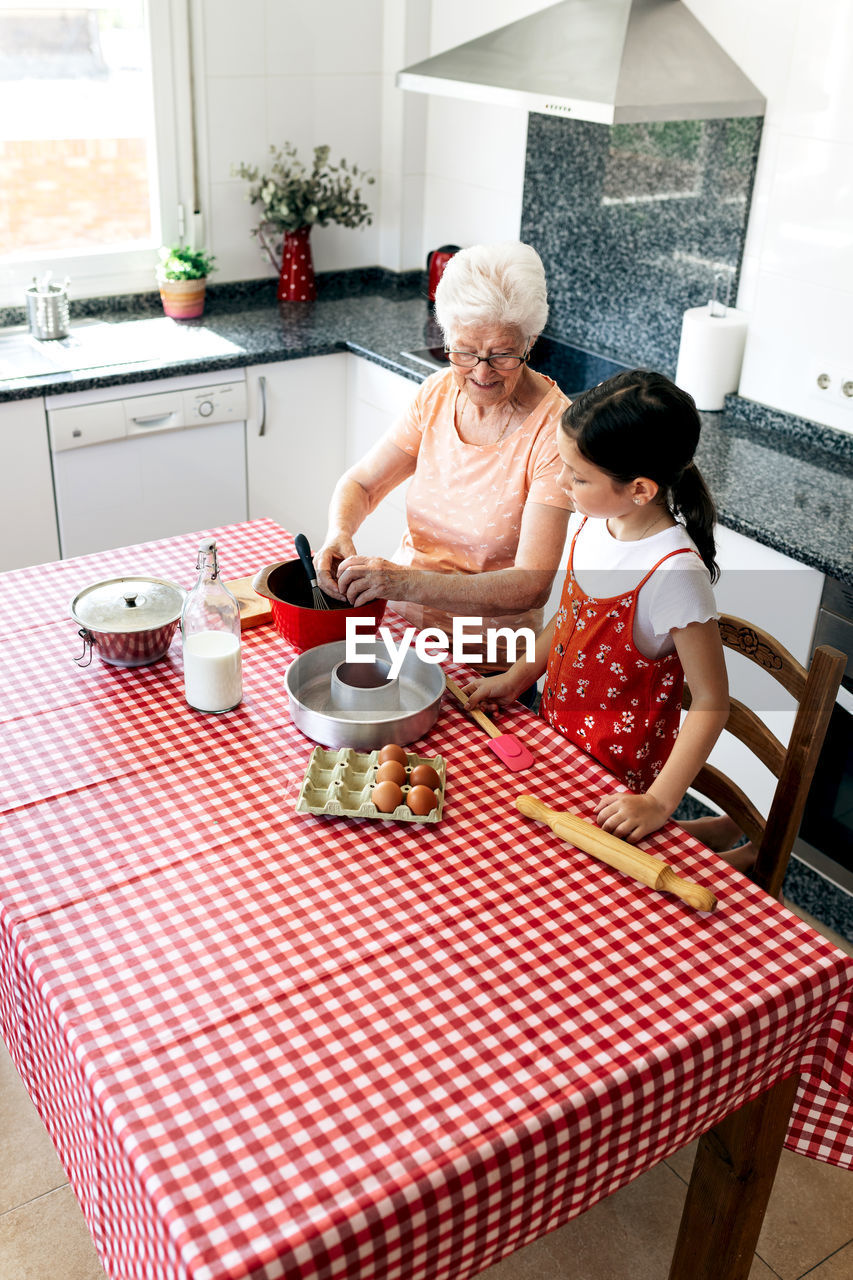 From above of grandma talking to girl at table with fresh ingredients and cake pan during cooking process at home