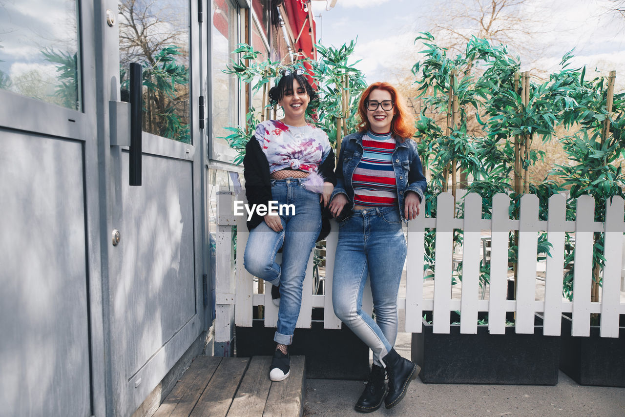 Portrait of hipster female friends standing against fence by building
