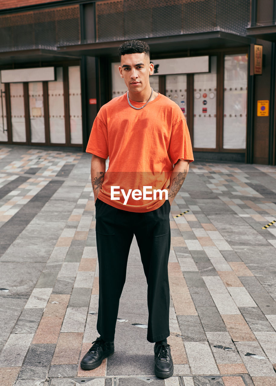 Portrait of young man standing on footpath against building