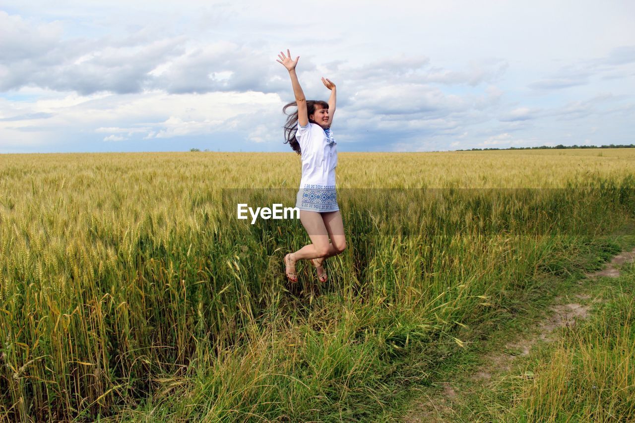 Cheerful young woman with arms raised jumping on field against cloudy sky