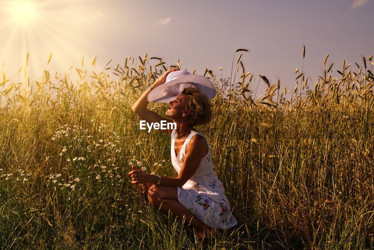 Side view of woman shielding eyes while hat while crouching amidst plants