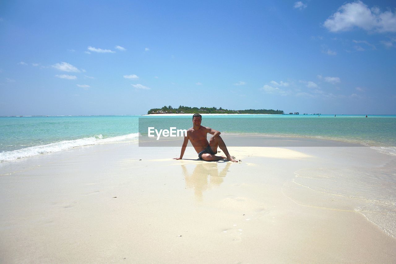 Full length portrait of shirtless man standing at beach against blue sky