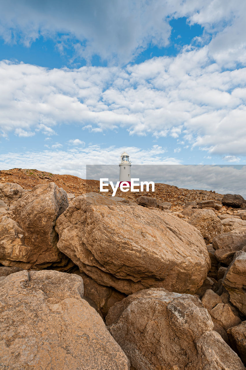 Tip of a lighthouse on rocks against cloudy sky.