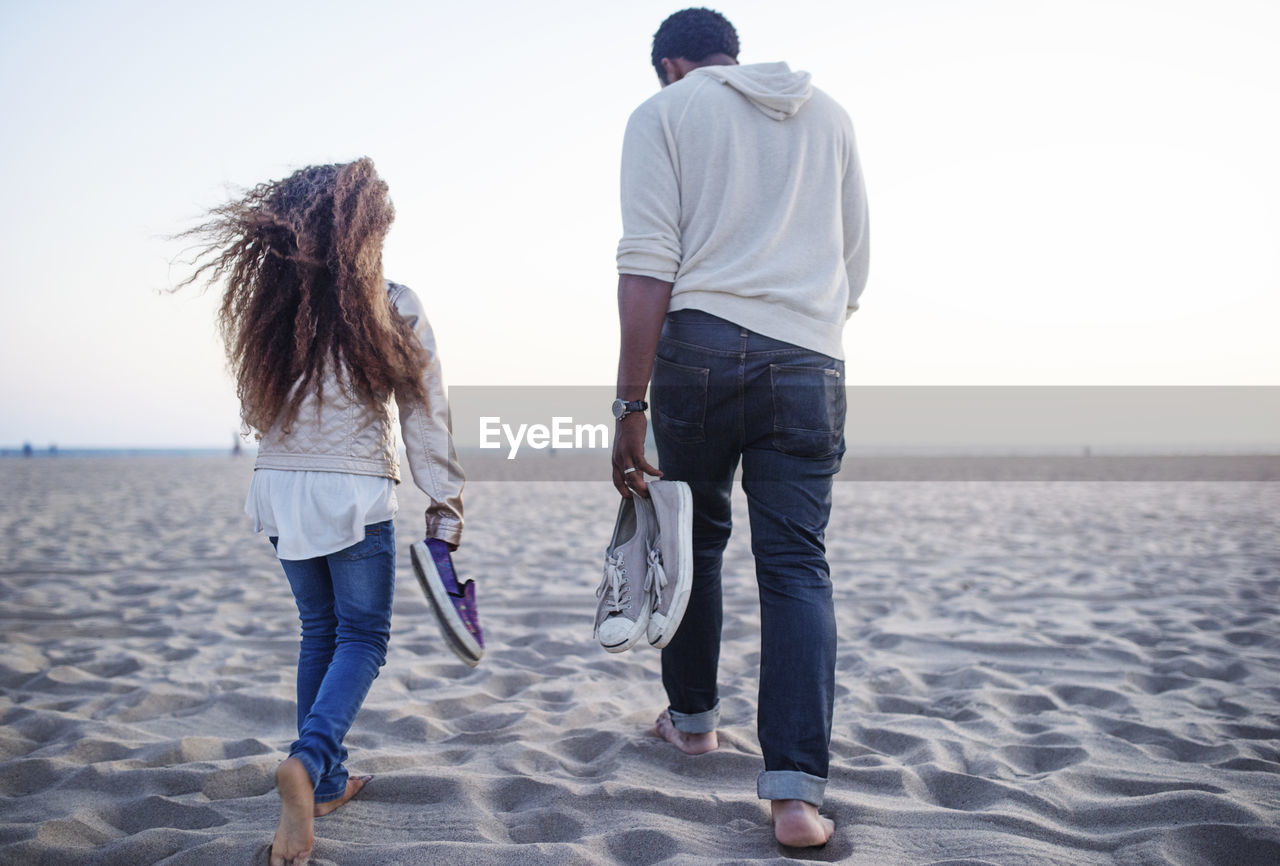 Rear view of father and daughter carrying shoes while walking at beach against clear sky