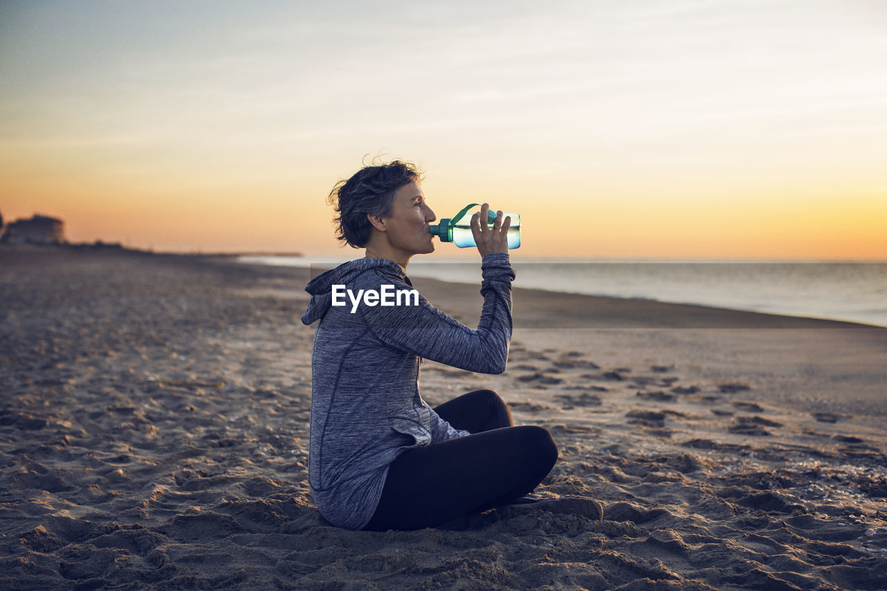 Woman drinking water while sitting at beach against sky