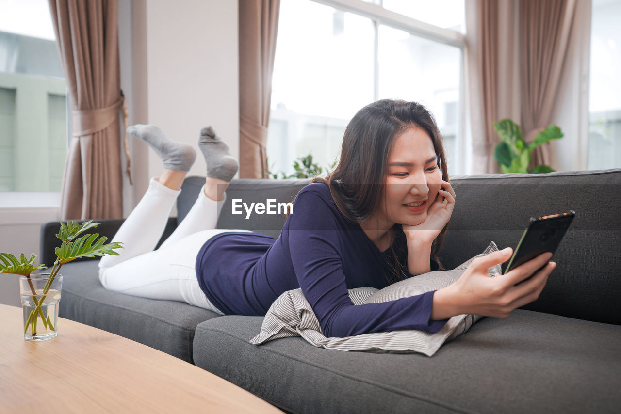 WOMAN USING MOBILE PHONE WHILE SITTING ON SOFA AT HOME