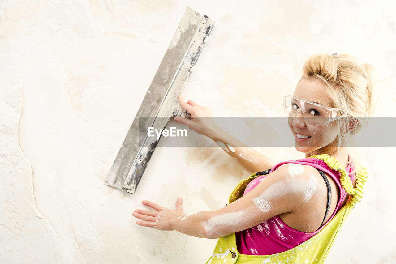 Portrait of young woman in coveralls working with putty knife against white wall