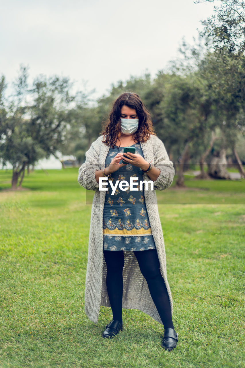 Full length of woman wearing mask holding phone standing at park