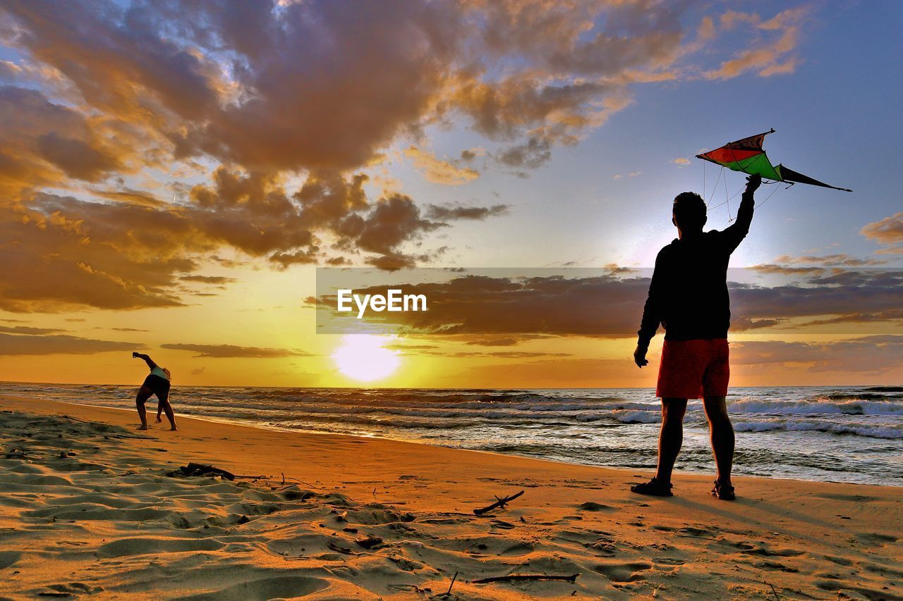 SILHOUETTE FRIENDS STANDING ON BEACH AGAINST SKY DURING SUNSET