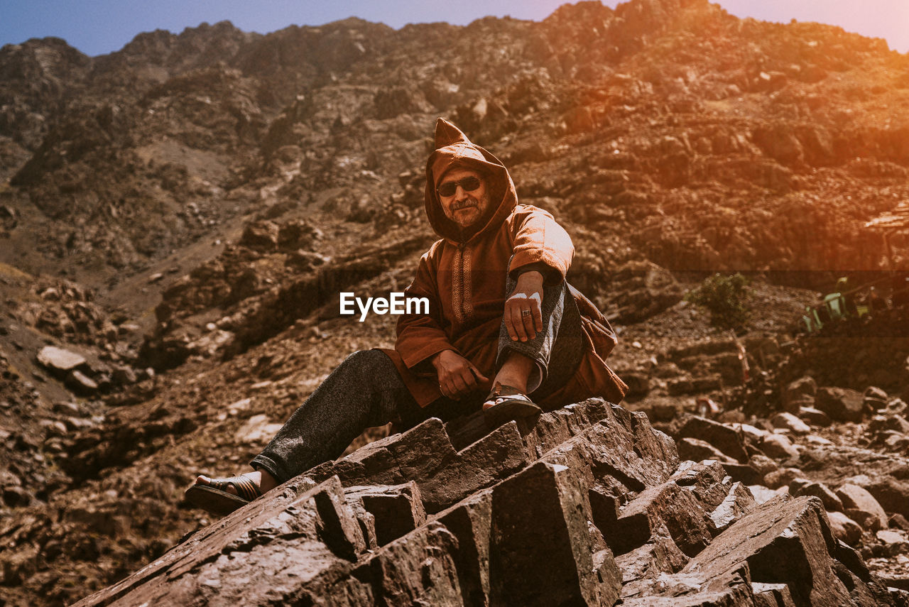 PORTRAIT OF YOUNG MAN SITTING ON ROCK AGAINST MOUNTAINS