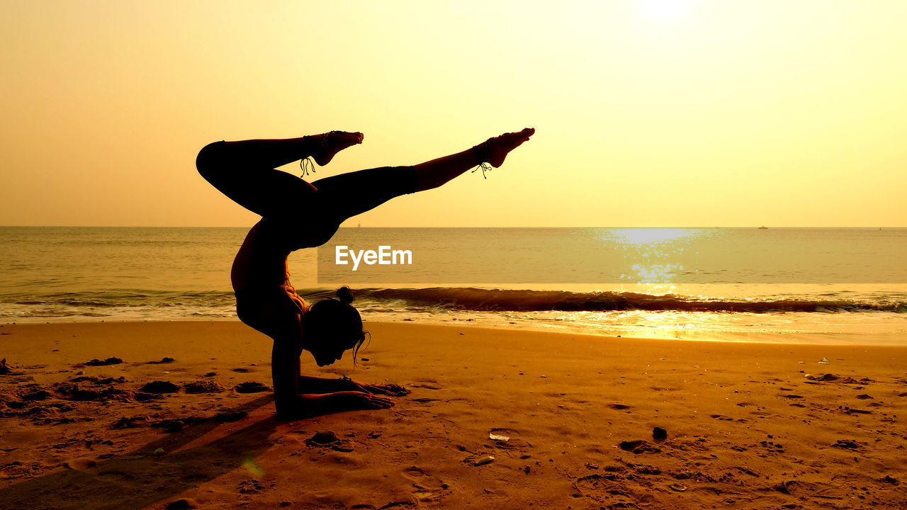 Full length of woman practicing yoga on shore at beach against sky during sunset