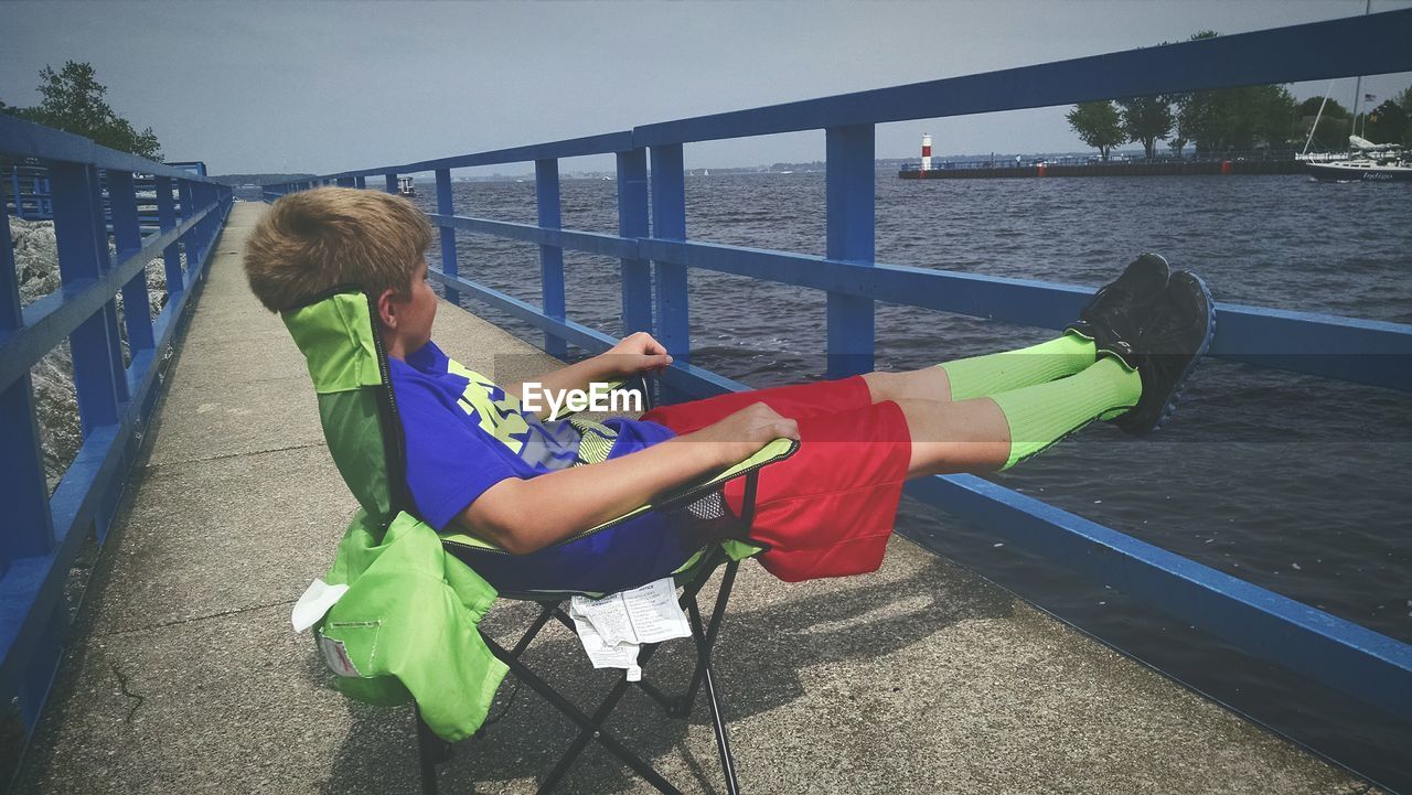 Side view of boy relaxing on chair by sea on walkway