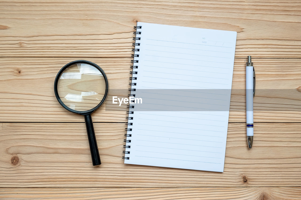 Directly above shot of spiral notebook with pen and magnifying glass on wooden table