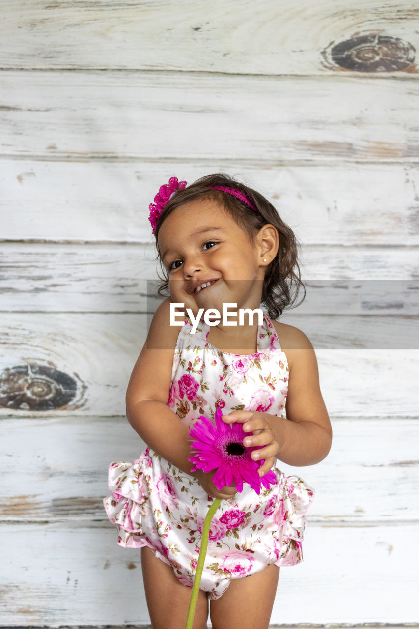 Smiling girl holding pink flower against wooden wall