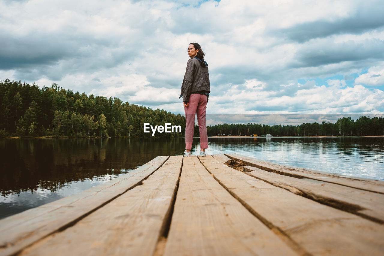 The tourist girl stands with her back on the wooden pier of the forest lake and turns around