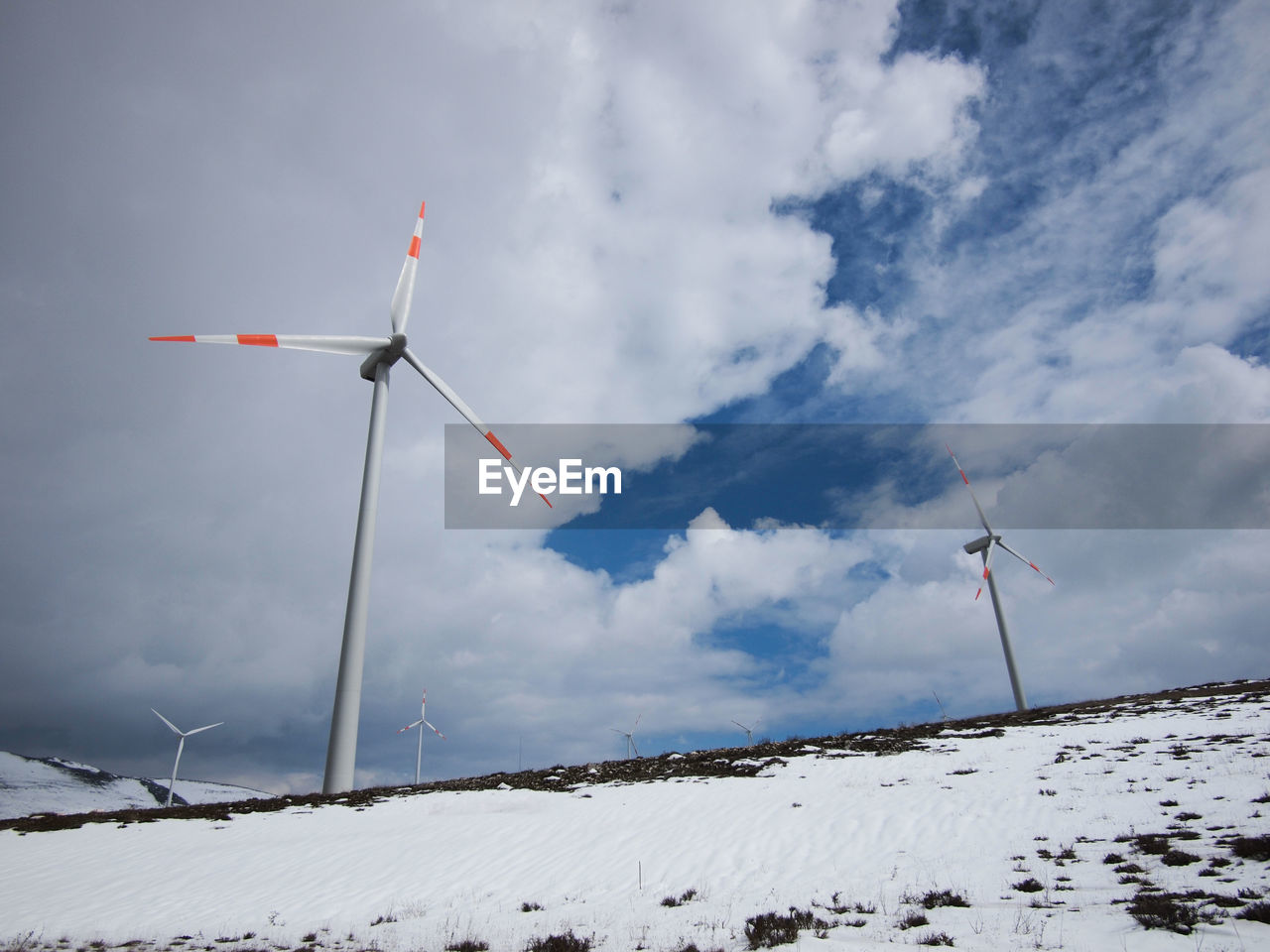 Low angle view of wind turbines on snowy field against cloudy sky