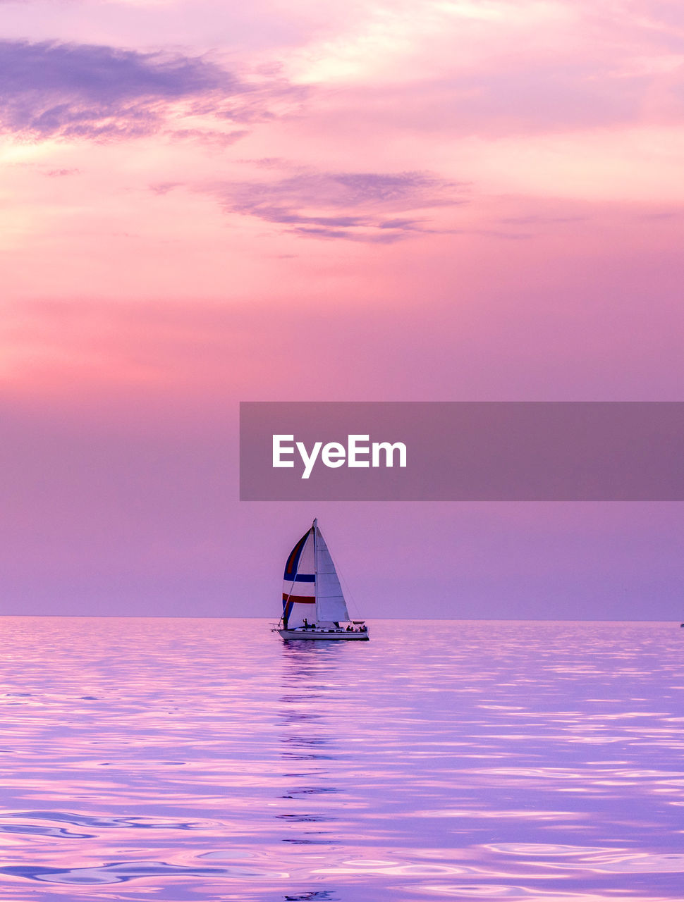 A lone sail boat floats on a still lake with a beautiful sunset of purple and pink